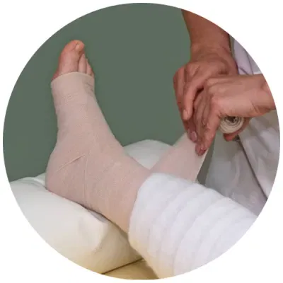 A close-up image of a foot being bandaged for lymphoedema treatment. A therapist is applying compression bandages to the ankle and foot, which are resting on a padded surface.